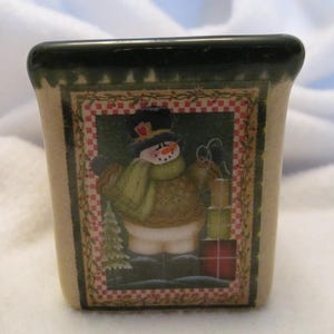 May include: A ceramic square-shaped container with a green rim and a festive snowman design. The snowman is wearing a green scarf and hat and holding a red gift box. The background is a red and white checkered pattern.