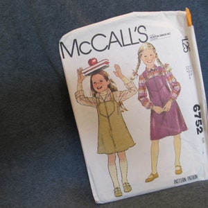 May include: McCall's sewing pattern number 6752 for a girl's dress. The pattern shows two girls wearing different styles of dresses. One dress is a purple jumper with a plaid shirt and the other is a brown jumper with a white shirt. The pattern is priced at $1.25.