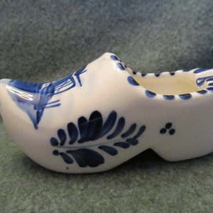 May include: A white ceramic Dutch shoe with blue and white Delftware designs. The shoe has a small opening at the top, suggesting it may be an ashtray.