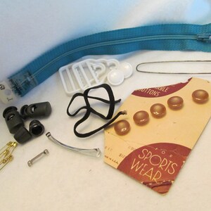 May include: Assortment of sewing and craft supplies including a turquoise zipper, white plastic buckles, black elastic, brown buttons, safety pins, and metal clasps. The label reads "For Sports Wear."