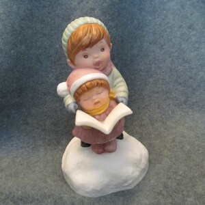 May include: A ceramic figurine depicting two children singing carols. One child, wearing a pink hat and coat, is asleep while holding a songbook. The other child, wearing a green hat and coat, is singing. The figurine is on a white base.