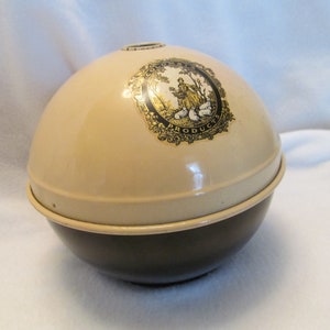 May include: A two-tone, spherical container with a beige upper half and a dark brown lower half. A gold and black emblem is affixed to the top, featuring a pastoral scene. The container appears to be made of metal.