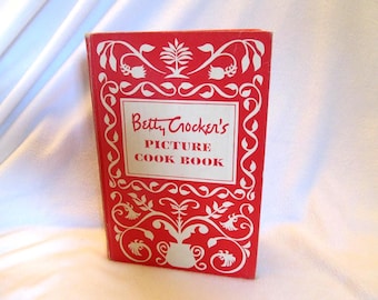 May include: A vintage cookbook titled "Betty Crocker's Picture Cook Book." The book has a red cover with white floral and scroll designs. The title is in a white rectangular box. The book is standing upright.