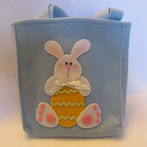 May include: A light blue felt Easter basket with a white bunny holding a yellow and green Easter egg. The bunny has pink ears and feet.
