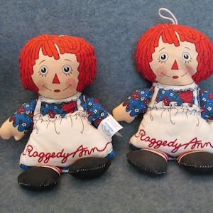 May include: Two Raggedy Ann dolls with red yarn hair, painted faces, and blue floral dresses. Each doll wears a white apron with "Raggedy Ann" embroidered in red and a heart that says "Love". Black shoes complete the look.