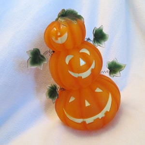 May include: A stack of three orange plastic jack-o'-lantern decorations with painted-on faces. Each pumpkin has a green leafy stem and is decorated with green plastic leaves.