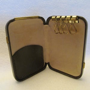 May include: A brown leather key holder with a flap that opens to reveal a key ring with five metal loops. The key holder has a small pocket on the inside of the flap.