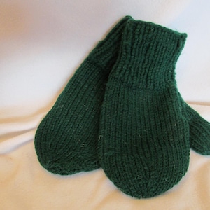 Puede incluir: Un par de mitones de punto verde oscuro. Los mitones están hechos de un hilo grueso y tienen un puño acanalado. Los mitones están sobre una superficie blanca. Un accesorio de invierno.