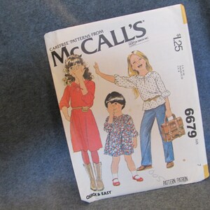 May include: A McCall's sewing pattern for girls' dresses. The pattern is number 6679 and costs $1.25. The pattern shows three girls wearing different dresses. The girl on the left is wearing a red dress with a white collar and a brown belt. The girl in the middle is wearing a blue and white floral dress. The girl on the right is wearing a white dress with a blue and white polka dot pattern.