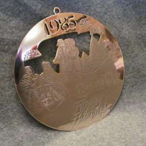 May include: A gold-toned Christmas ornament with a cut-out design of a winter scene. The ornament features the year "1985" and the text "Happy Holidays" and "From Our Family To Yours".