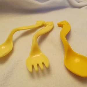 May include: Three yellow plastic baby utensils: two spoons and a fork. The handles are curved and have a decorative floral design at the top. The utensils are on a white surface.