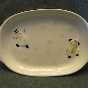 May include: Oval, off-white ceramic serving platter with raised handles. The platter features a snowman and a snow-person design, along with embossed snowflakes. The snowman has a black top hat and coal eyes. The snow-person has a red nose and green accents.
