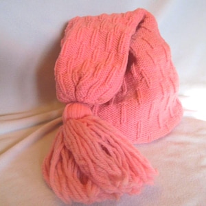 Puede incluir: Un gorro de punto de color coral con un diseño largo y plegado y una borla grande. El gorro presenta un patrón texturizado y está hecho de hilo suave. La borla está hecha del mismo hilo de color coral.