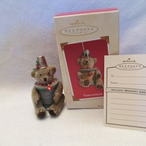 May include: A brown teddy bear wearing a thimble on its head is sitting inside a spool of thread. The bear is holding a small red heart. The ornament is in its original box, which is red and white and says "Hallmark Keepsake Ornament" on the front. The box also includes a tag that says "Holiday Memory 2002".