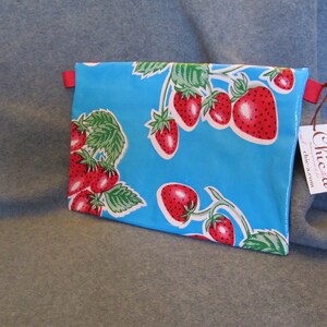 Puede incluir: Una cartera de mano de tela azul con una cremallera roja y una correa roja. La bolsa est&aacute; decorada con un patr&oacute;n de fresas rojas sobre un fondo azul. La bolsa tiene una etiqueta blanca adjunta con el texto "Chica chic-a-rum".
