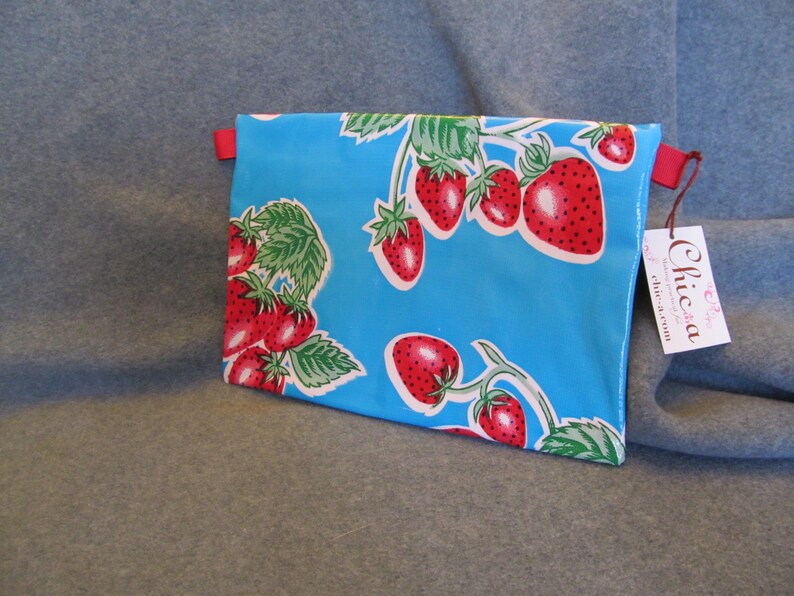 Puede incluir: Una cartera de mano de tela azul con una cremallera roja y una correa roja. La bolsa est&aacute; decorada con un patr&oacute;n de fresas rojas sobre un fondo azul. La bolsa tiene una etiqueta blanca adjunta con el texto "Chica chic-a-rum".
