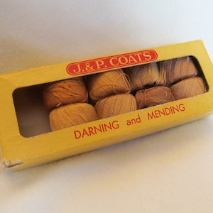 May include: A vintage box of J. & P. Coats darning and mending thread. The yellow box has a rectangular window revealing several spools of tan thread. The text "J. & P. COATS" is in a red oval, and "DARNING and MENDING" is printed in red.