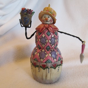 May include: A snowman ornament with a pink and purple patchwork dress, a straw hat, and a small basket of flowers in one hand. The snowman is holding a small pink and gray trowel in the other hand.