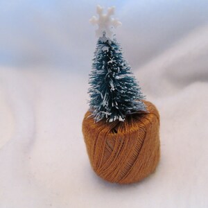 May include: A miniature Christmas tree with a white snowflake topper sits atop a ball of brown yarn. The tree has green needles and is dusted with white. The yarn ball is a warm, earthy tone, creating a festive, handcrafted feel.