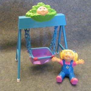 May include: A blue and purple swing set with a cabbage patch kid doll sitting on the swing. The swing set has a green cabbage patch kid head on top.