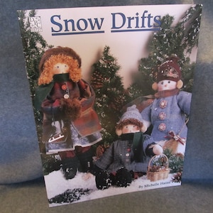 May include: A book titled "Snow Drifts" by Michelle Haines, featuring a cover illustration of three handmade dolls dressed in winter clothing. The dolls are standing in a snowy scene with pine trees.