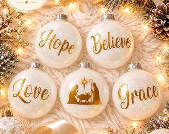May include: Five round white glittery Christmas ornaments featuring gold lettering and imagery. The ornaments display the words Hope, Believe, Love, and Grace in a cursive font, while the center ornament depicts a gold nativity scene silhouette.