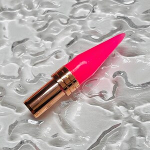 May include: A vibrant pink lipstick with a gold-coloured metal base and a pointed tip. The lipstick is set against a textured, silvery-white background, creating a striking contrast.