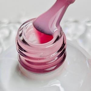 May include: A clear glass bottle with a pink nail polish inside. A pink brush is dipping into the polish.