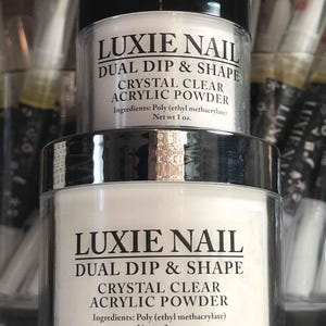 Könnte beinhalten: Zwei transparente Gläser mit Luxie Nail Acrylpulver. Die Gläser haben schwarze Deckel und silberne Ränder. Auf den Etiketten steht "DUAL DIP & SHAPE CRYSTAL CLEAR ACRYLIC POWDER". Ein Glas enthält 28g, das andere 57g Pulver.