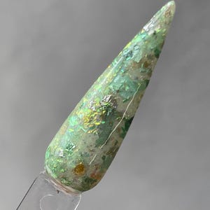 May include: A close-up of a green nail polish with gold and silver glitter. The polish is on a clear nail tip.