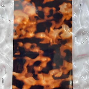 May include: A close-up of a sheet of brown and orange tortoiseshell patterned material. The material has a glossy finish and a swirling pattern.