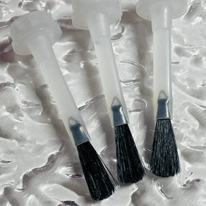 May include: Three clear plastic nail polish applicator brushes with black bristles. The brushes are arranged diagonally on a textured white surface. The applicators have a clear plastic handle and a cap.