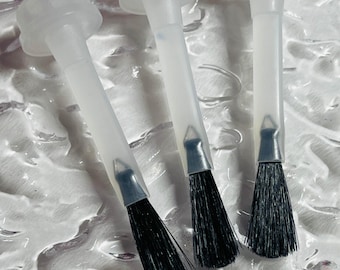 May include: Three clear plastic nail polish applicator brushes with black bristles. The brushes are arranged diagonally on a textured white surface. The applicators have a clear plastic handle and a cap.