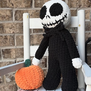 Peut inclure: Une grande poupée au crochet faite à la main ressemblant à Jack Skellington, avec une tête blanche et des traits noirs, portant un costume noir. Une citrouille orange avec une tige verte est à côté de la poupée.