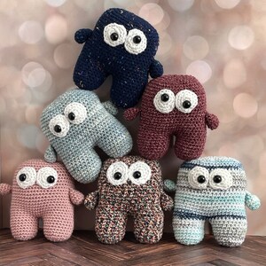 May include: Six crocheted plush toys in different colors and patterns, each with large white eyes and black pupils. The toys are shaped like a simple humanoid figure with two arms and two legs.