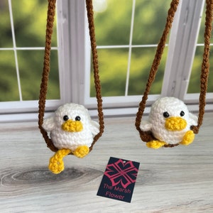 May include: Two white crocheted birds with yellow beaks and feet are hanging from brown cords. The birds are sitting on a wooden surface. The text "The Marsh Flower" is visible on a tag below the birds.