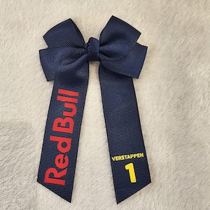 May include: A navy blue hair bow with a red "Red Bull" logo on one side and a yellow "1" with "VERSTAPPEN" text on the other side.