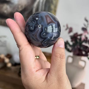 May include: A polished, spherical stone featuring intricate, swirling patterns in shades of dark gray, charcoal, and translucent white. The surface is smooth and reflective, displaying natural banding and organic, cloud-like formations throughout the orb.
