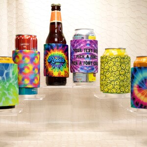 Op de afbeelding: Een assortiment drankisolatoren, elk met een uniek ontwerp. Stijlen zijn onder meer tie-dye, smiley-gezichten en een ontwerp met het woord "Relax". De isolatoren zijn gewikkeld rond een bierfles en diverse blikjes.