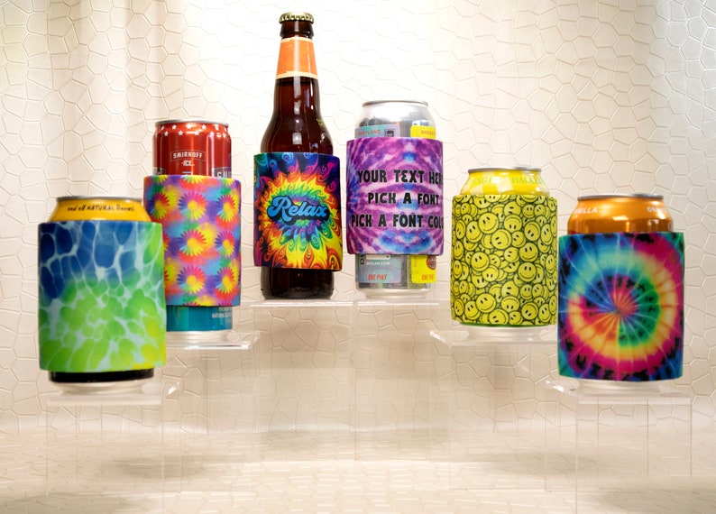 Op de afbeelding: Een assortiment drankisolatoren, elk met een uniek ontwerp. Stijlen zijn onder meer tie-dye, smiley-gezichten en een ontwerp met het woord "Relax". De isolatoren zijn gewikkeld rond een bierfles en diverse blikjes.
