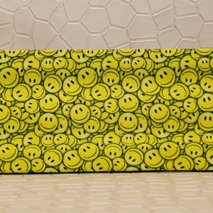 Op de afbeelding: Rechthoekige brillenkoker met een herhaald patroon van gele smileygezichten op een zwarte achtergrond. De koker heeft afgeronde hoeken en een gladde textuur, wat zorgt voor een vrolijk ontwerp. De koker is ontworpen om brillen te beschermen.