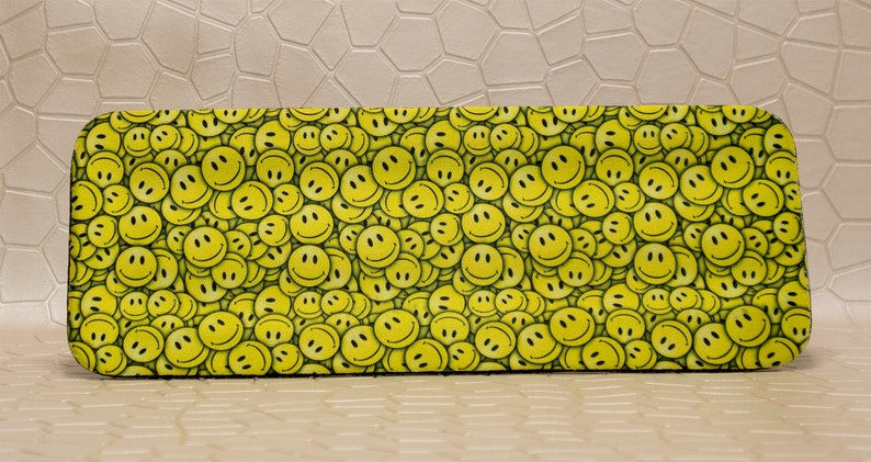 Op de afbeelding: Rechthoekige brillenkoker met een herhaald patroon van gele smileygezichten op een zwarte achtergrond. De koker heeft afgeronde hoeken en een gladde textuur, wat zorgt voor een vrolijk ontwerp. De koker is ontworpen om brillen te beschermen.