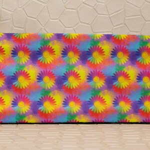 Op de afbeelding: Een rechthoekige, kleurrijke muismat met een herhalend patroon van madeliefachtige bloemen. De bloemen hebben gele centra en bloemblaadjes in roze, oranje en paarse tinten, tegen een achtergrond in aquarel-stijl.