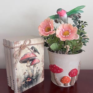 Puede incluir: Una pila de tres libros atados con cordel y un botón de madera, con una ilustración de setas y un pájaro con el texto NATURE BEAUTY LOVE, junto a una maceta de metal beige con flores rosas, follaje verde y una figura de colibrí.
