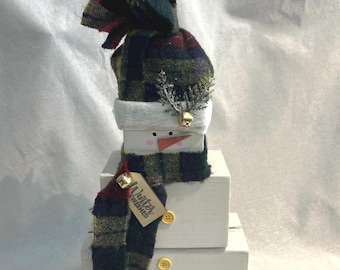 Puede incluir: Decoración de muñeco de nieve artesanal hecha de bloques de madera blancos apilados. Presenta un sombrero y una bufanda a cuadros en tonos verdes, rojos y azul marino. El muñeco de nieve tiene una nariz de zanahoria, ojos de botón y una etiqueta que dice "Winter Wishes".