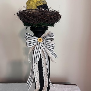 Könnte beinhalten: Dekorative schwarze Säule mit einem Vogel in einem Nest. Der Vogel ist gelb, schwarz und weiß. Die Säule ist mit einer schwarz-weiß gestreiften Schleife und einem cremefarbenen Spitzenband verziert, mit einem Holzknopf in der Mitte.