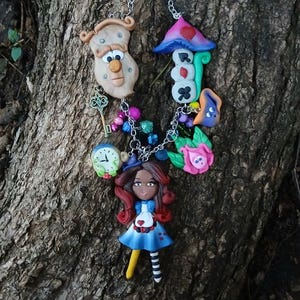 May include: A whimsical necklace featuring Alice in Wonderland-themed charms. The necklace includes a miniature Alice figure, a clock, a key, a mushroom, and other colorful elements. The charms are connected by a silver chain.