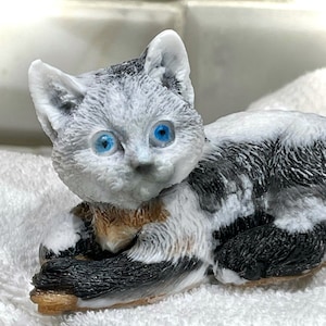May include: A white and black cat figurine with blue eyes, lying on a white fluffy towel. The cat is made of soap and has a realistic appearance.