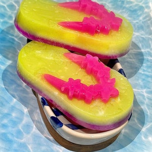 May include: Two yellow and pink layered soap bars shaped like flip flops with pink flower designs. The soap bars are sitting on a white and blue dish with a wooden base. The text "www.soapscentdesigns.com" is at the bottom of the image.