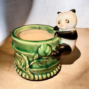 Puede incluir: Una maceta de cerámica con forma de tallo de bambú verde con una figura de panda tridimensional adherida al lateral. El panda es blanco con orejas, manchas en los ojos y extremidades negras, sobre una base de bambú con esmalte verde brillante.